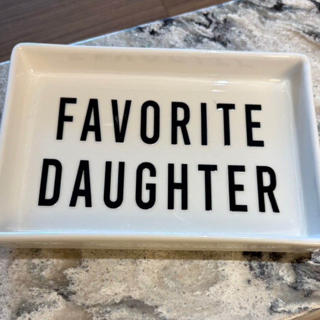 A rectangular white ceramic dish with the words 'FAVORITE DAUGHTER' printed in bold black capital letters, resting on a speckled granite countertop.