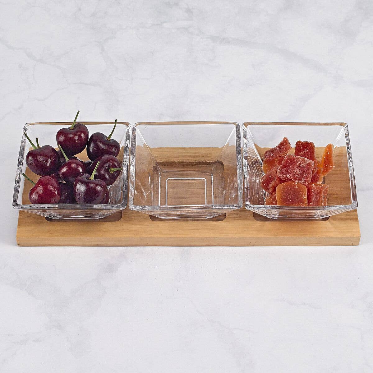 Three glass bowls on a wooden stand with cherries, a clear item, and dried fruits against a light background