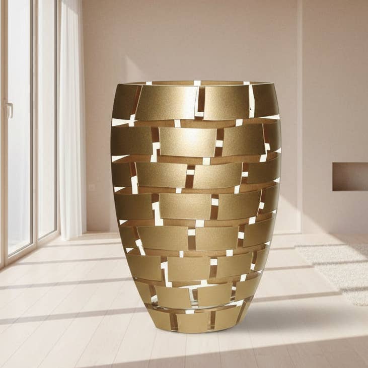 A modern cylindrical metal planter with a woven basket-like cutout pattern, finished in a brushed gold finish, set in a minimalist beige-toned room.