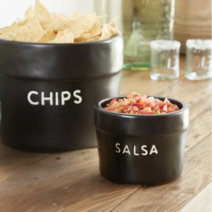 A set of two black ceramic bowls on a wooden table, labeled 'CHIPS' and 'SALSA' 