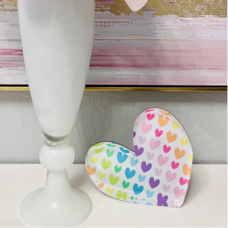 White vase with a pink flower on a white surface, next to a colorful heart-shaped object.